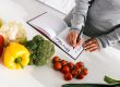 Mâini care notează un plan de mese într-un carnet, înconjurate de ardei, broccoli, conopidă și roșii