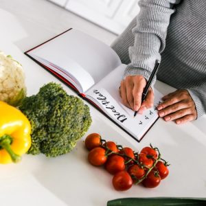 Cum citești corect eticheta unui aliment — 7 capcane de marketing 1 Mâini care notează un plan de mese într-un carnet, înconjurate de ardei, broccoli, conopidă și roșii