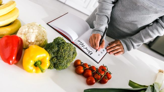 Mâini care notează un plan de mese într-un carnet, înconjurate de ardei, broccoli, conopidă și roșii