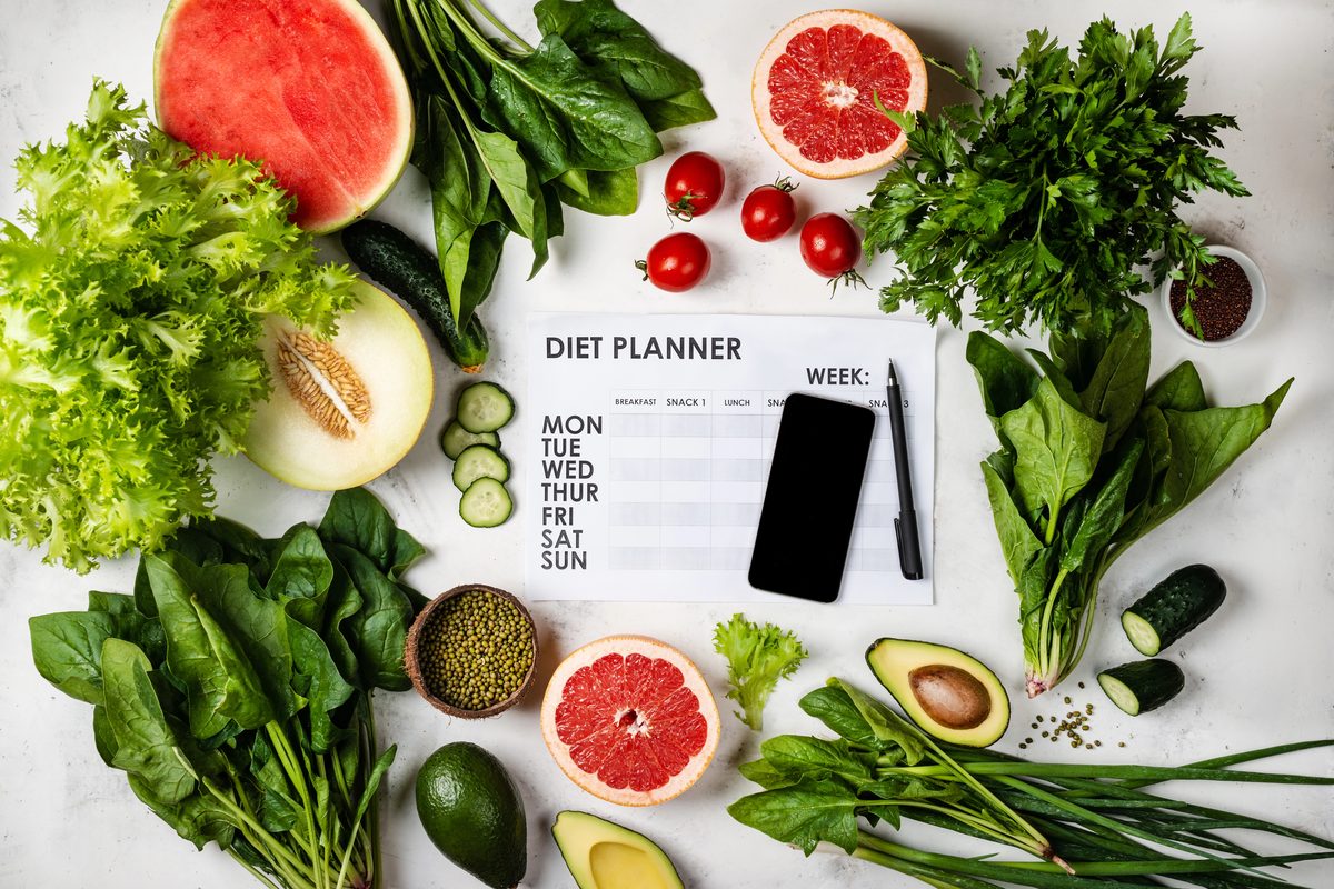 Cum citești corect eticheta unui aliment — 7 capcane de marketing 4 Un planificator alimentar săptămânal și un telefon, înconjurate de spanac, salată, avocado, pepene și grepfrut