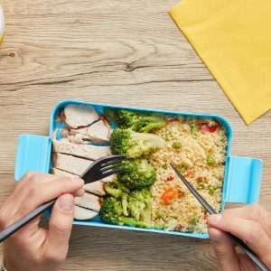 Pachet de prânz la birou: 12 combinații care nu se strică și nu te lasă flămând la 16:00 2 Persoană care mănâncă dintr-o cutie de prânz cu pui, broccoli și cuscus, la un birou de lemn, lângă o cafea